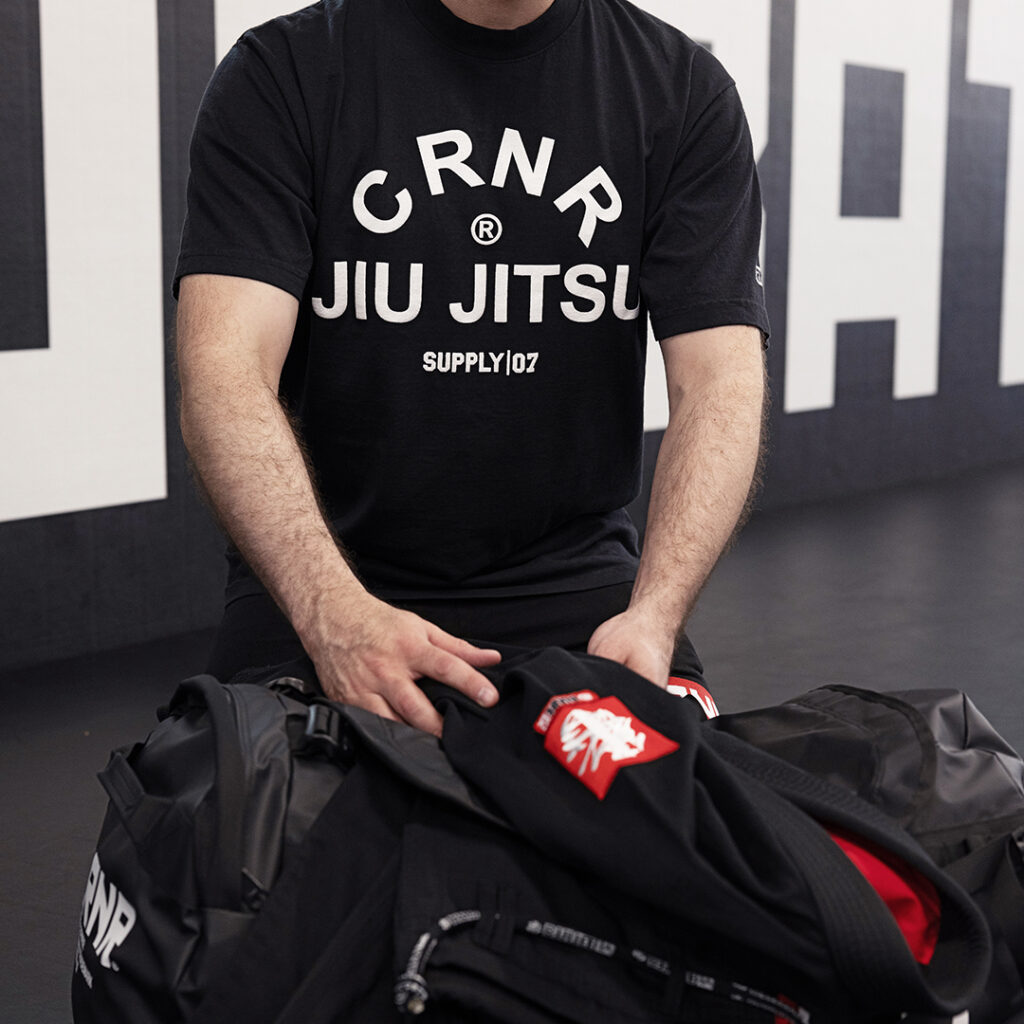 Photo of Combat Corner’s USA Made Essential Jiu Jitsu Combat Sport Tees. The photo shows a man in a Jiu Jitsu gym. The man is unpacking his gi and preparing for his Jiu Jitsu training session. We only see the man’s torso, and we see him wearing Combat Corner’s USA Made Essential Jiu Jitsu Combat Sport Tee. The essential tee is black, has a relaxed fit, and is made from USA cotton.