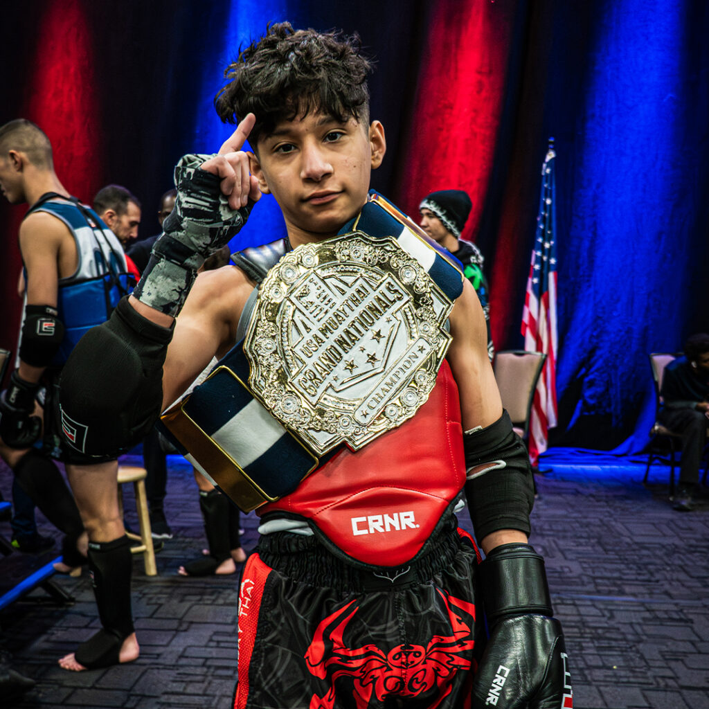 An photo of a youth Muay Thai fighter for Combat Corner's recap article of the 2026 USA Muay Thai Grand Nationals. The photo shows Muay Thai athletes competing at the tournament and using Combat Corner Muay Thai gear.
